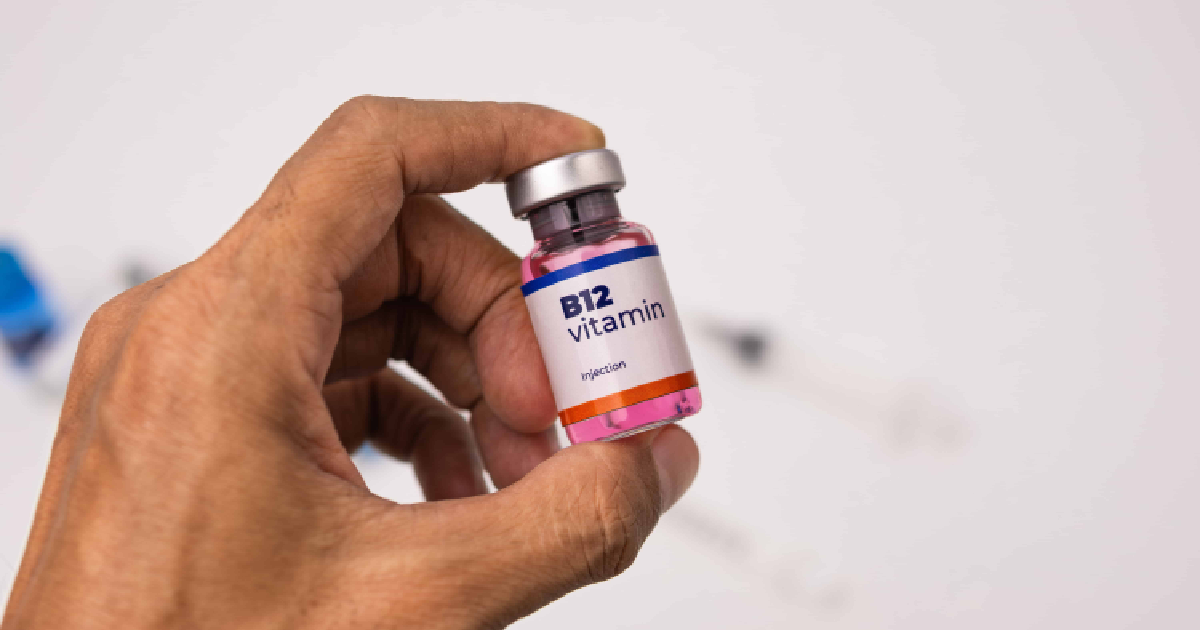 Hand holding a small pink B12 injections vial with a silver cap; a syringe is blurred in the background in Texas